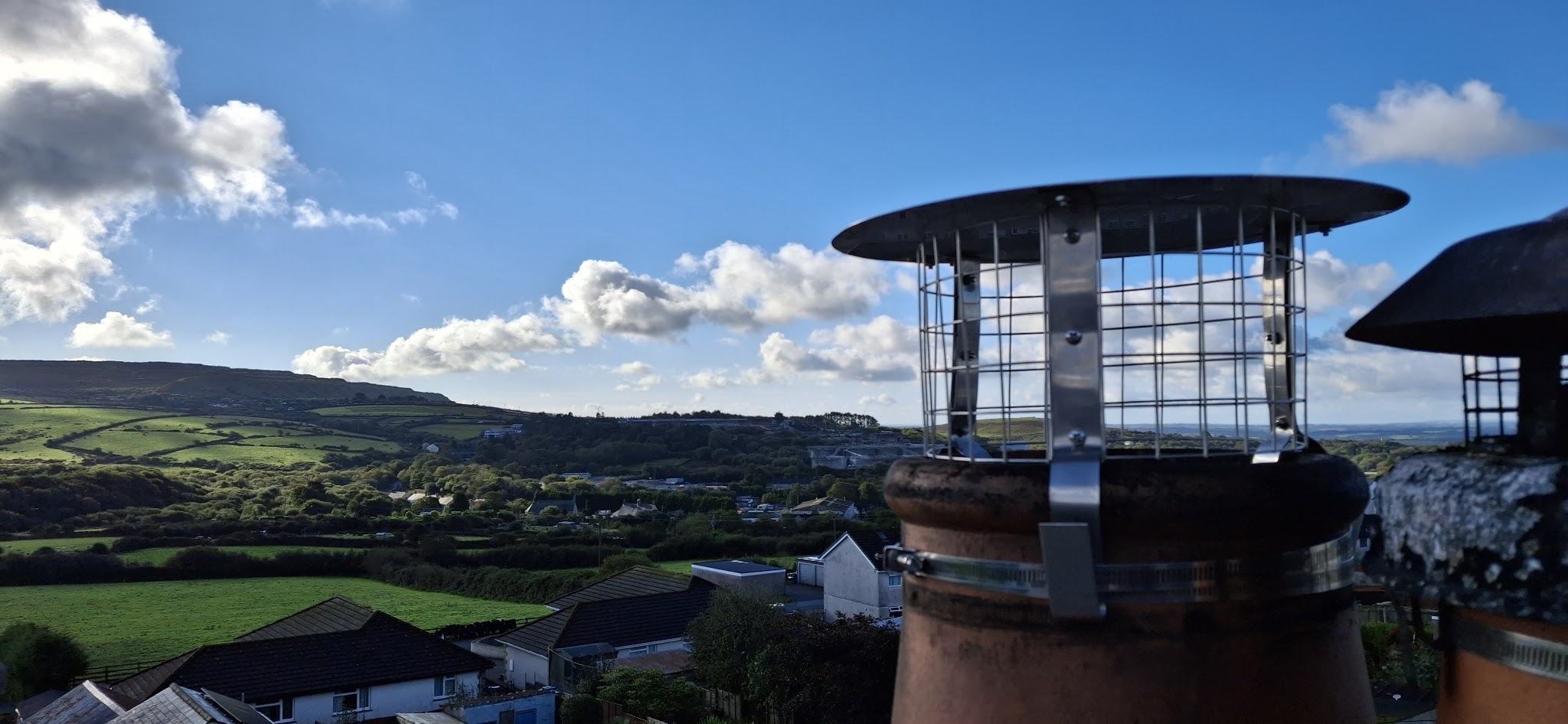 A stainless mesh cowl fitted on a chimney pot, with rolling Cornish countryside and the sea in the distance